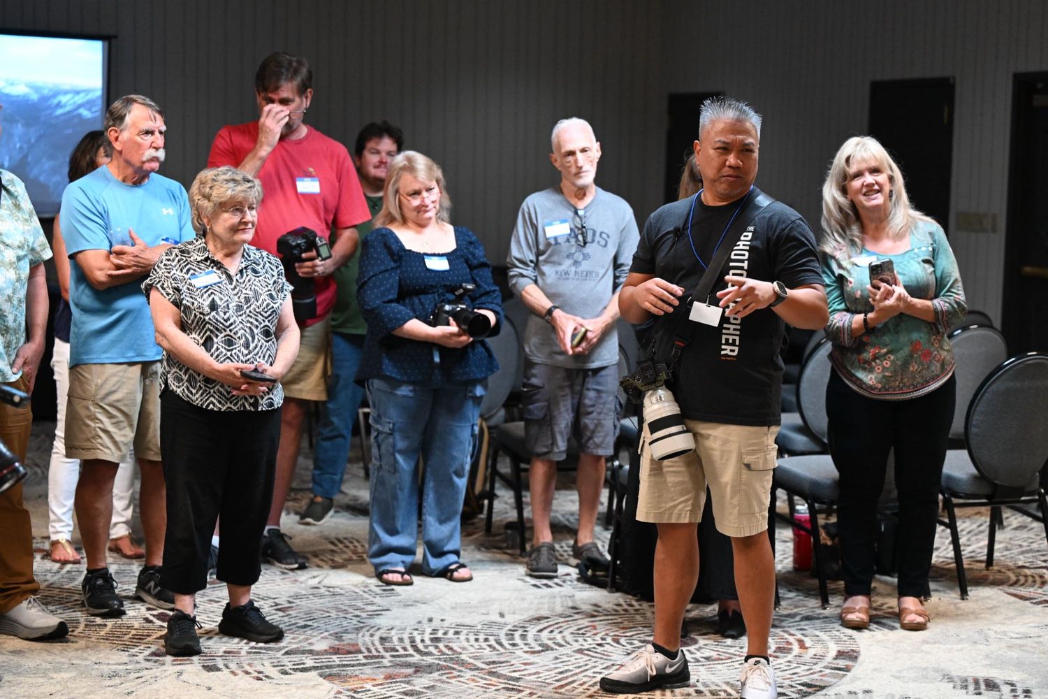 Group of adults gathered indoors at a photography club meeting, holding cameras on a patterned carpet floor.