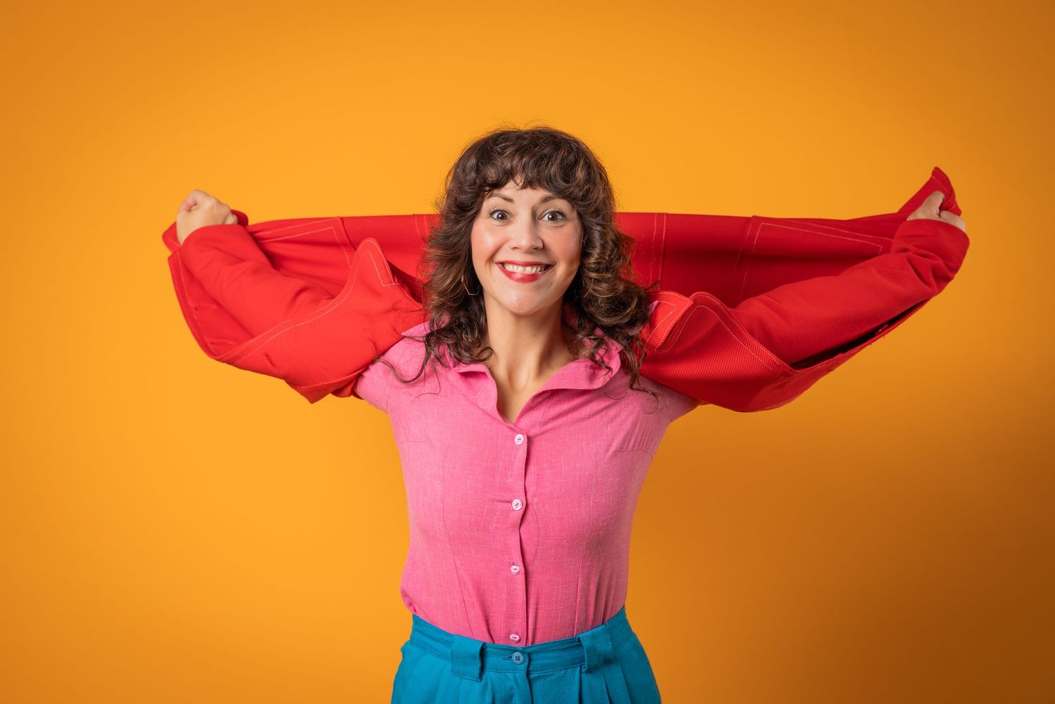 Woman in pink blouse holds red fabric wide against yellow background, smiling joyfully.