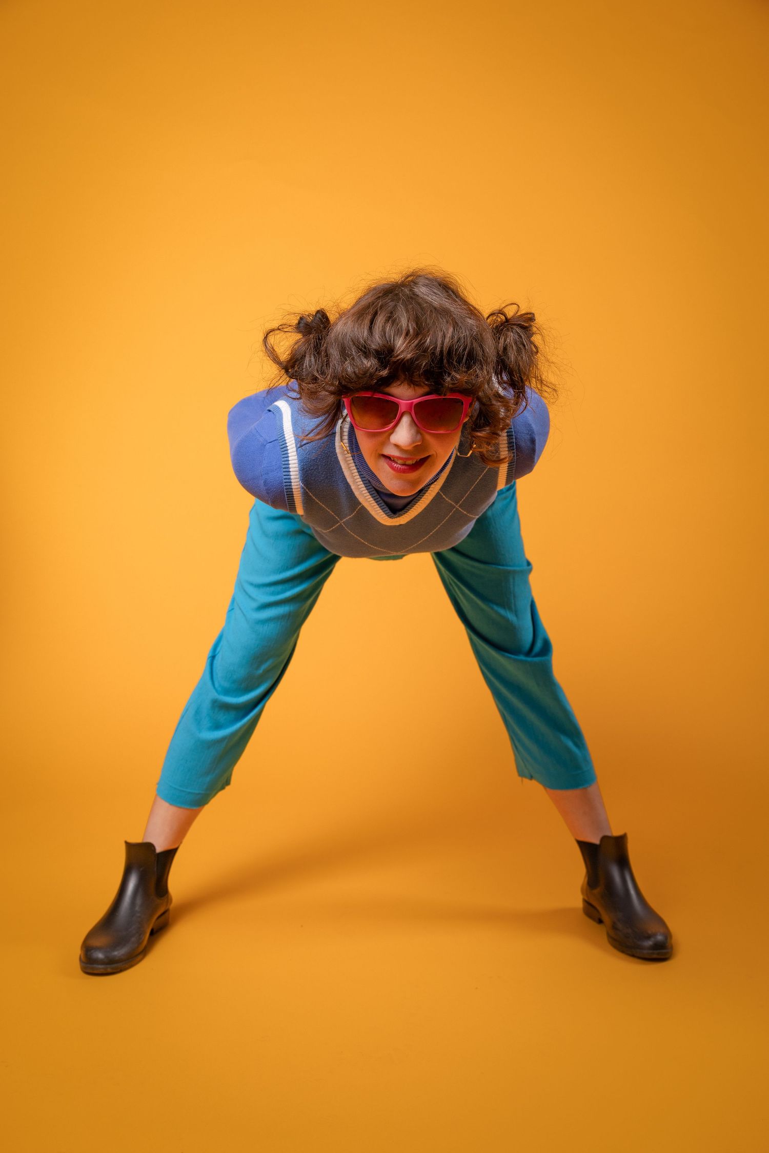 Funky woman in teal outfit and red heart sunglasses bending forward on yellow background.