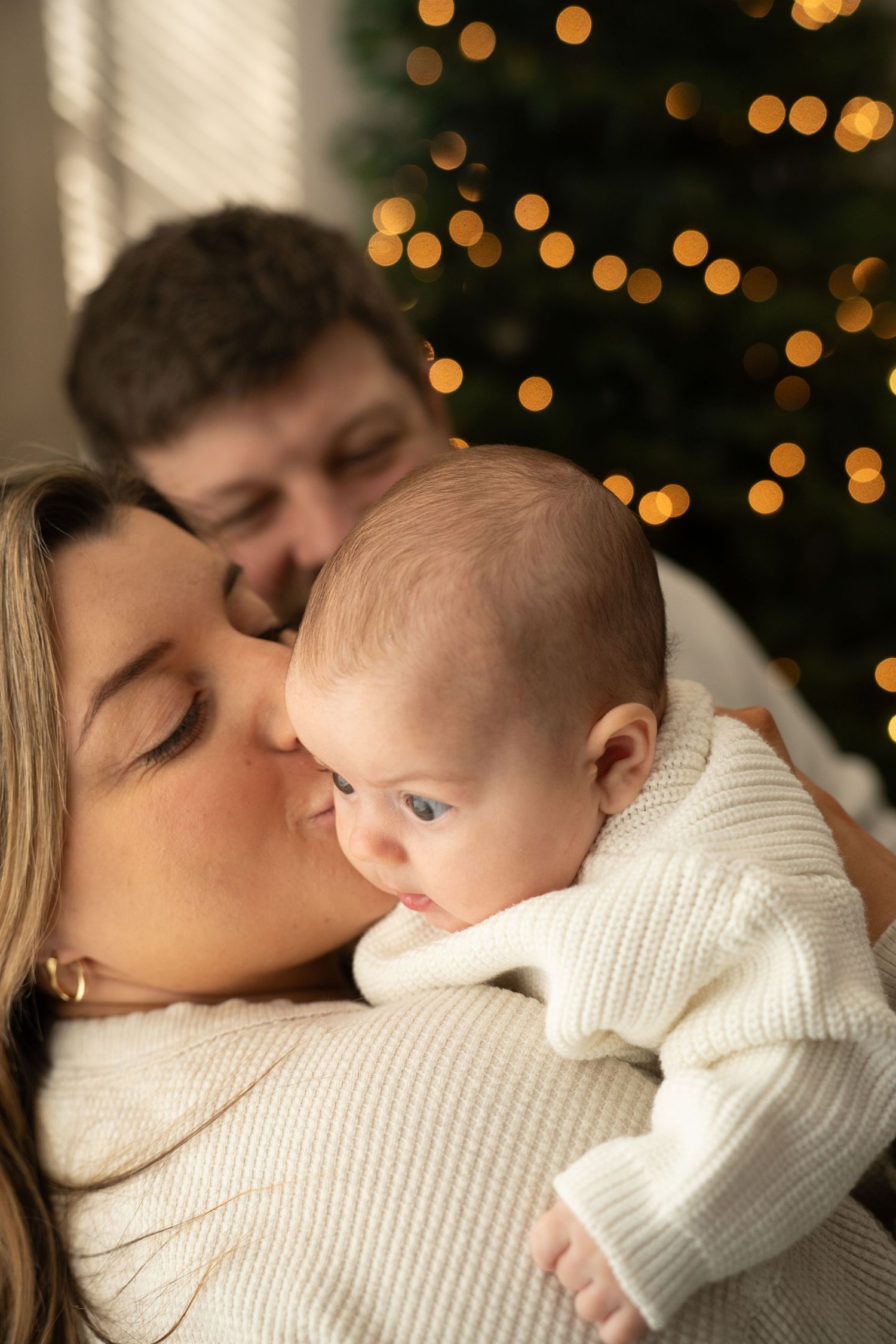 Cozy winter family moment with baby in white sweater against Christmas light bokeh.