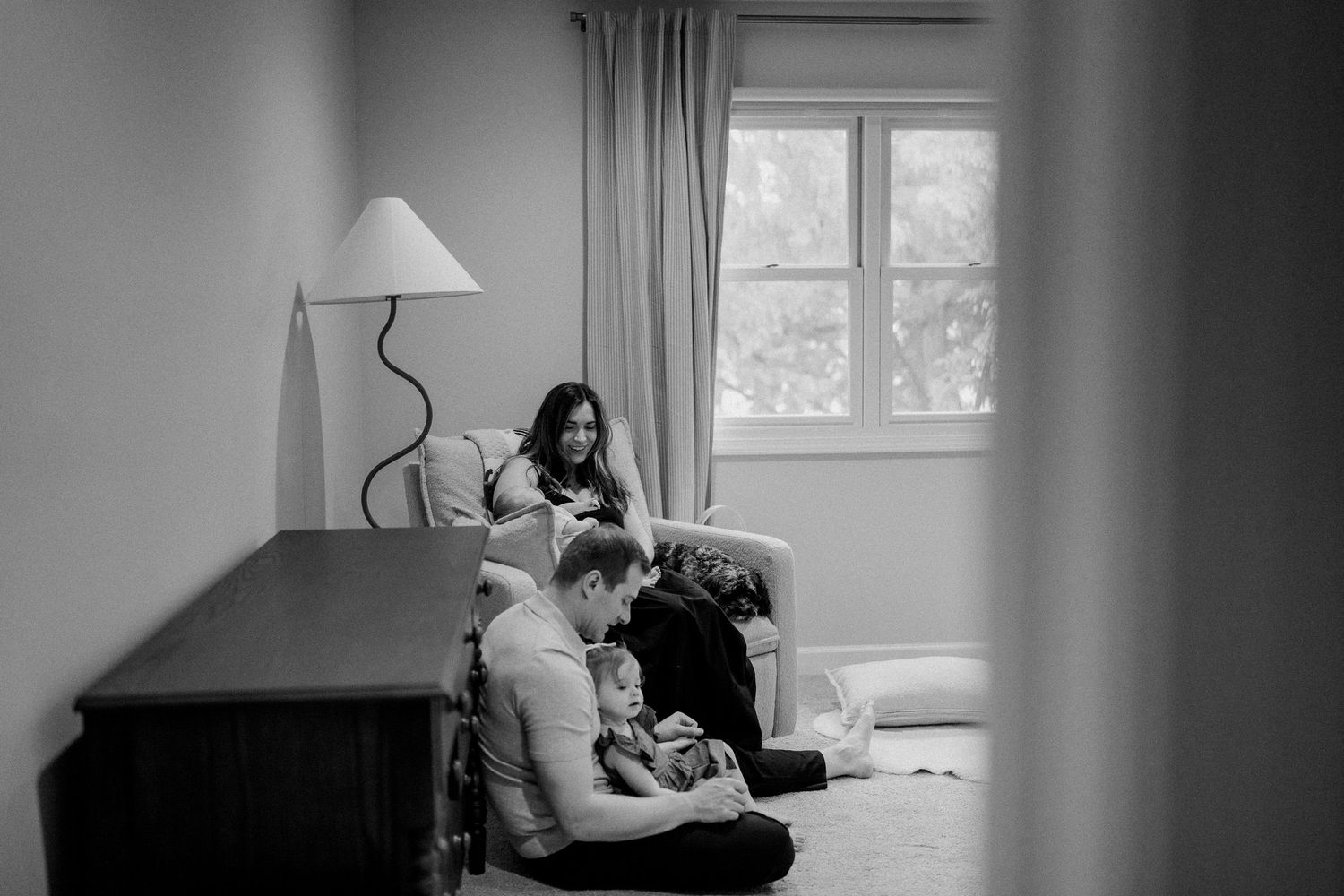 Black and white family photo of parents relaxing in a cozy room, one sitting on floor holding child, other reclining in chair.