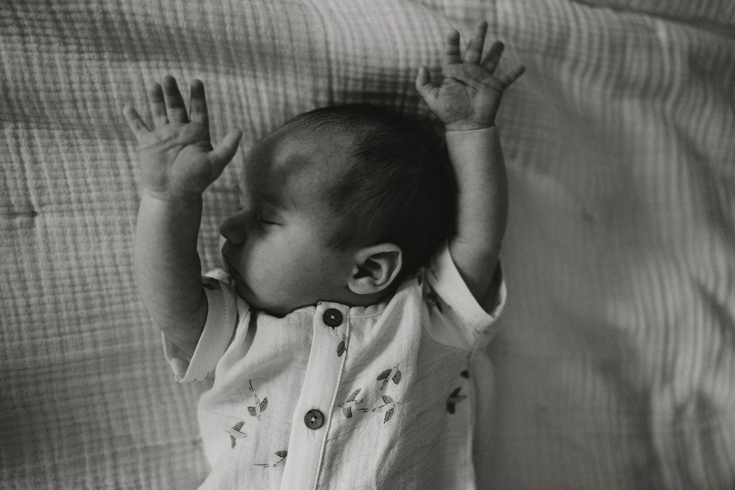 Black and white photo of a newborn baby lying on back with arms raised, wearing a white onesie.