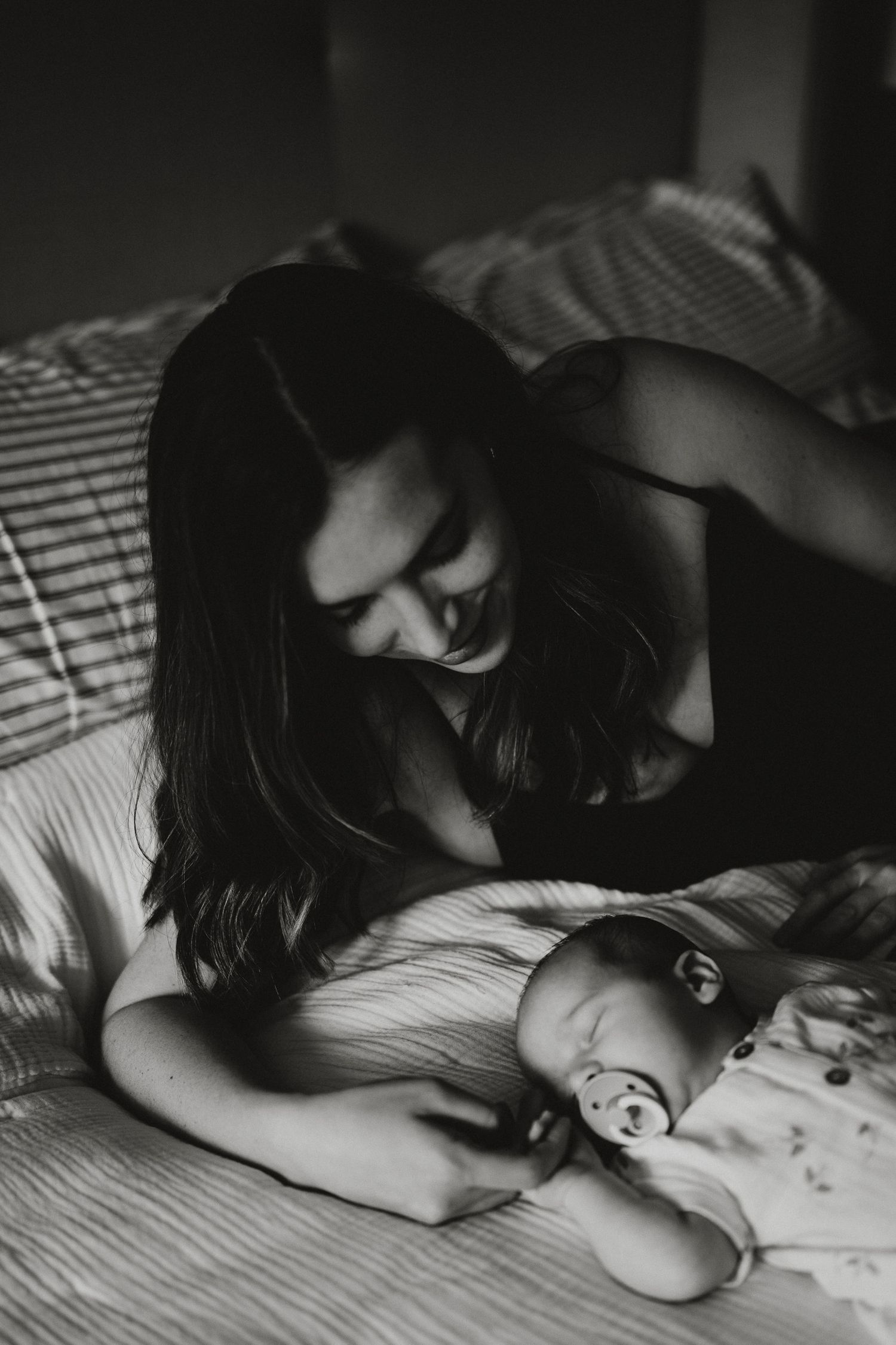 Black and white photo of a mother tenderly gazing at her newborn baby lying on a bed with a pacifier.