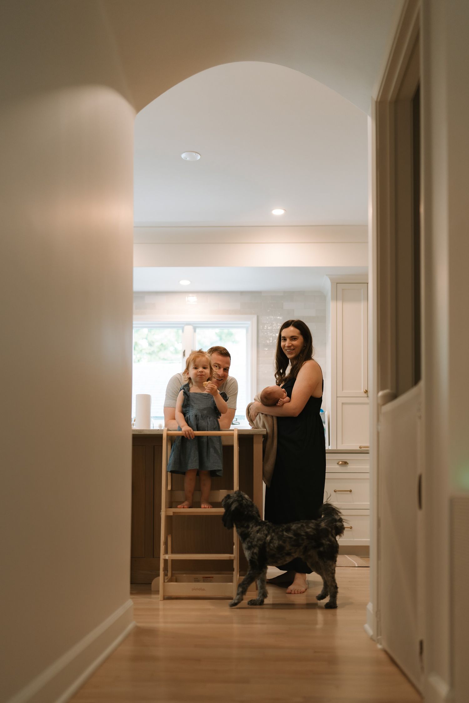 Woman in black dress holds newborn near two children on step stool and a dog in bright white kitchen.