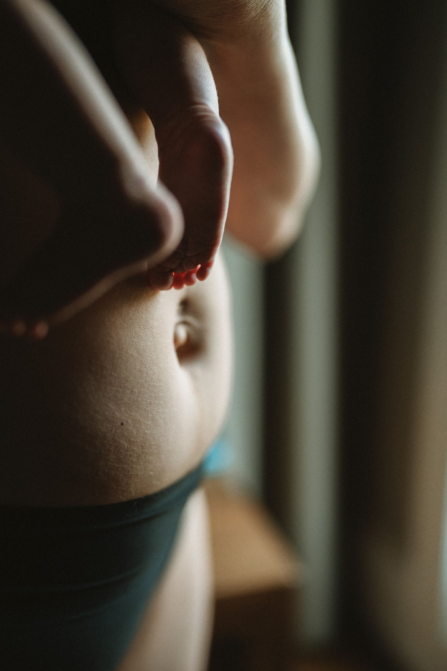 Close-up of a woman's bare midriff with a belly button piercing, wearing teal underwear, in soft natural light.
