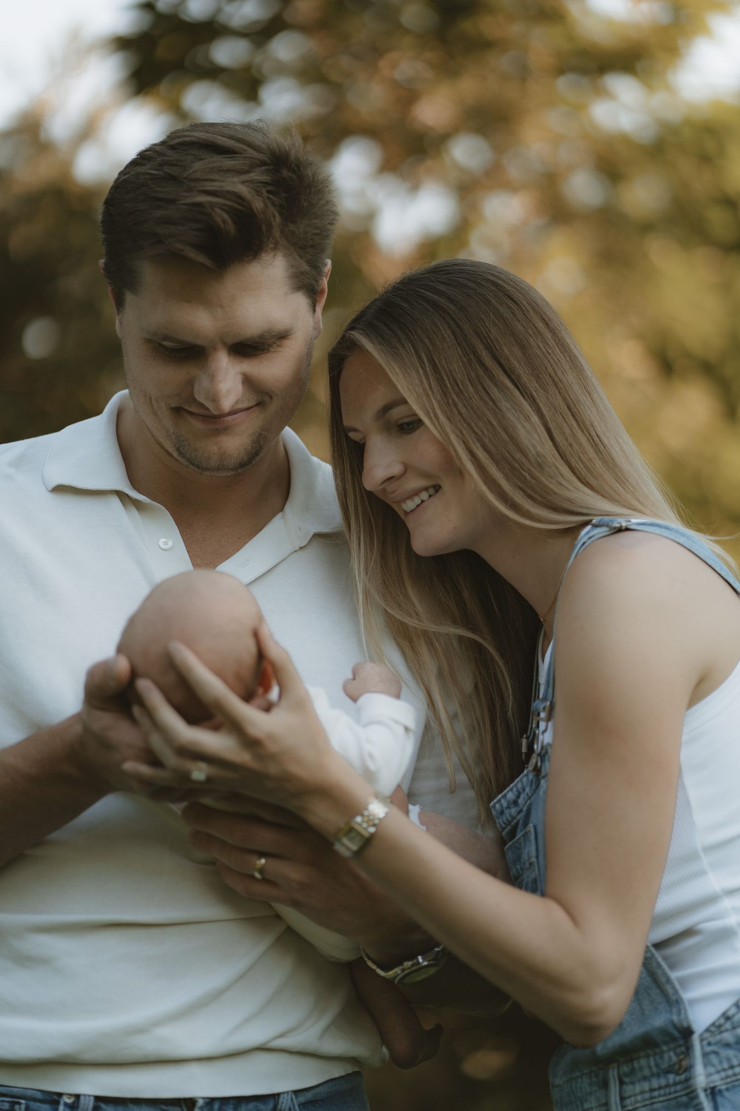 A young couple smiles lovingly at their newborn baby outdoors, surrounded by autumn foliage.