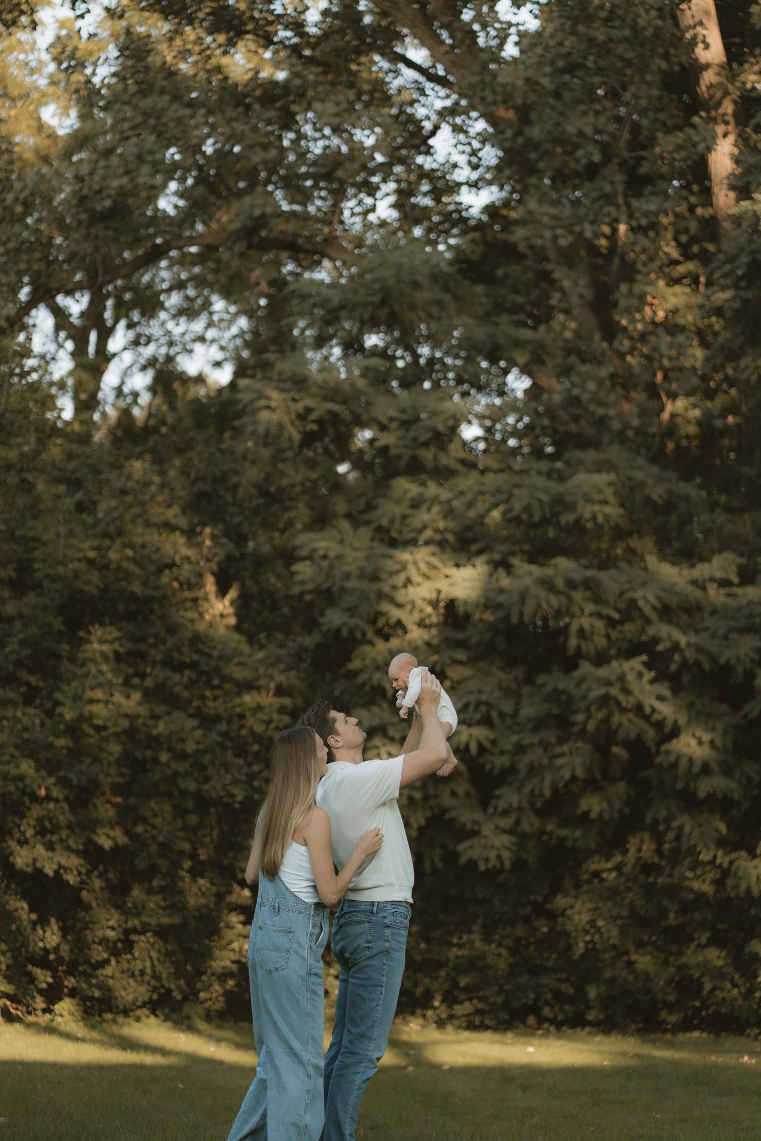 A couple stands on green grass lifting their baby in the air, surrounded by lush trees in warm golden light.