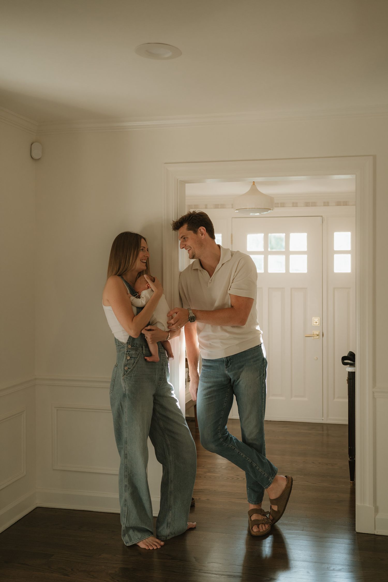 A young couple with a newborn baby stands in a bright home entryway, smiling at each other in casual jeans.