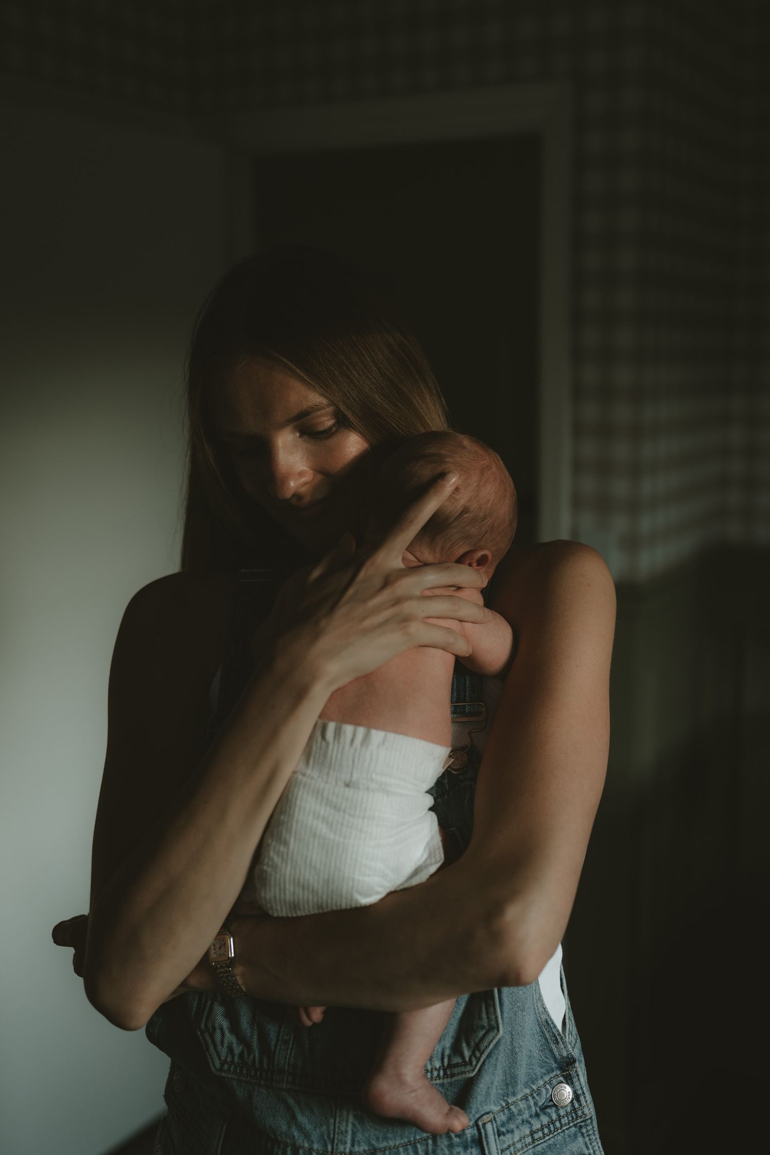 A young mother tenderly holds her newborn baby close in a softly lit, moody indoor setting.