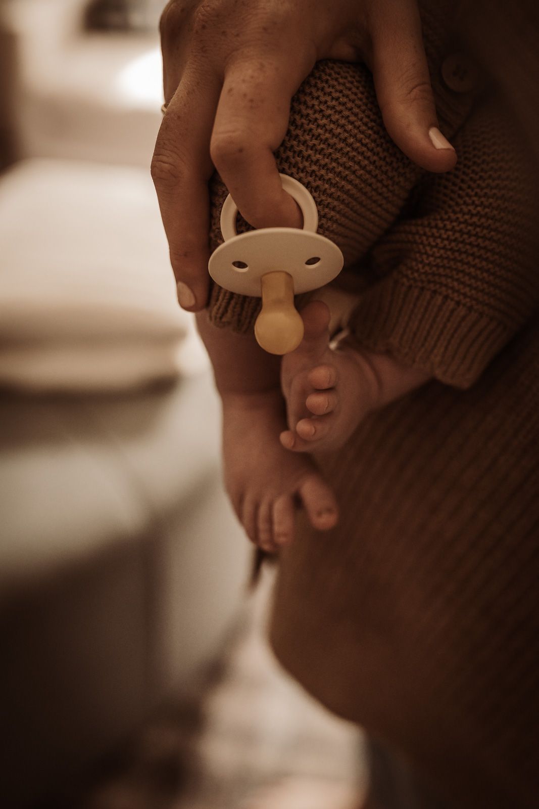 A parent gently holds a newborn baby's tiny feet alongside a pacifier, wrapped in a cozy knit sweater.