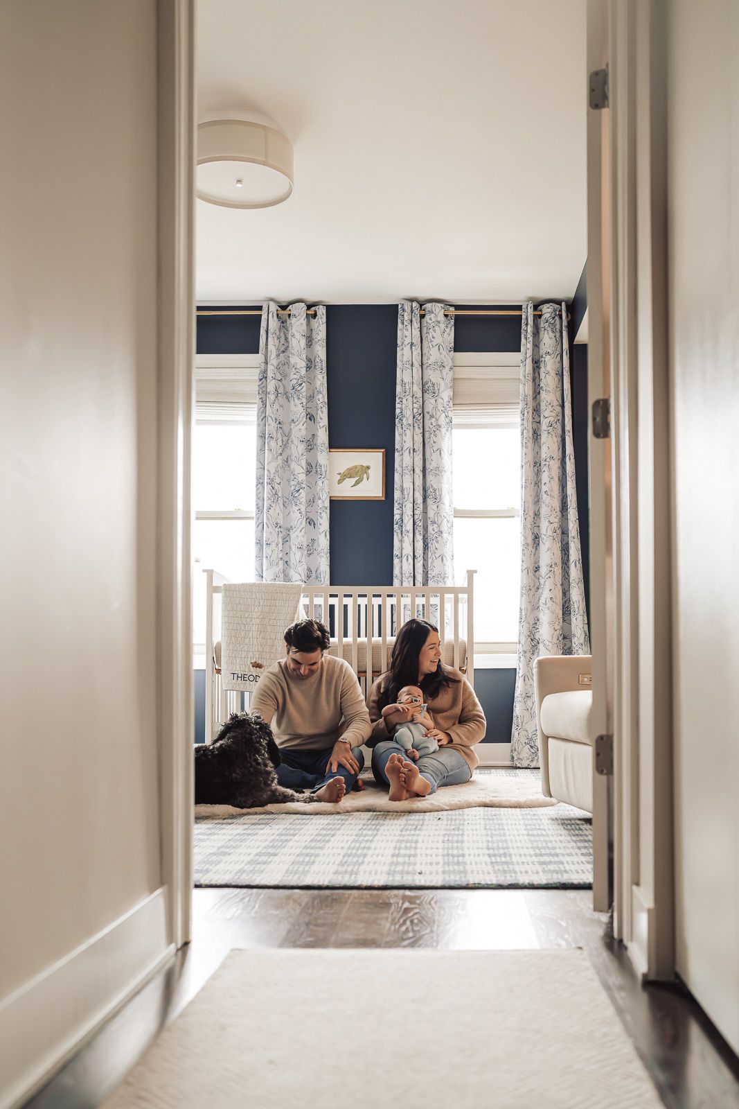 A young family sits on a rug in a navy blue nursery, playing with their baby and dog near a white crib.