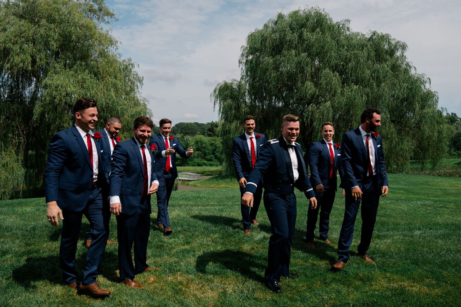 Groomsmen in navy suits gather for group photos on lawn with willow trees in background.
