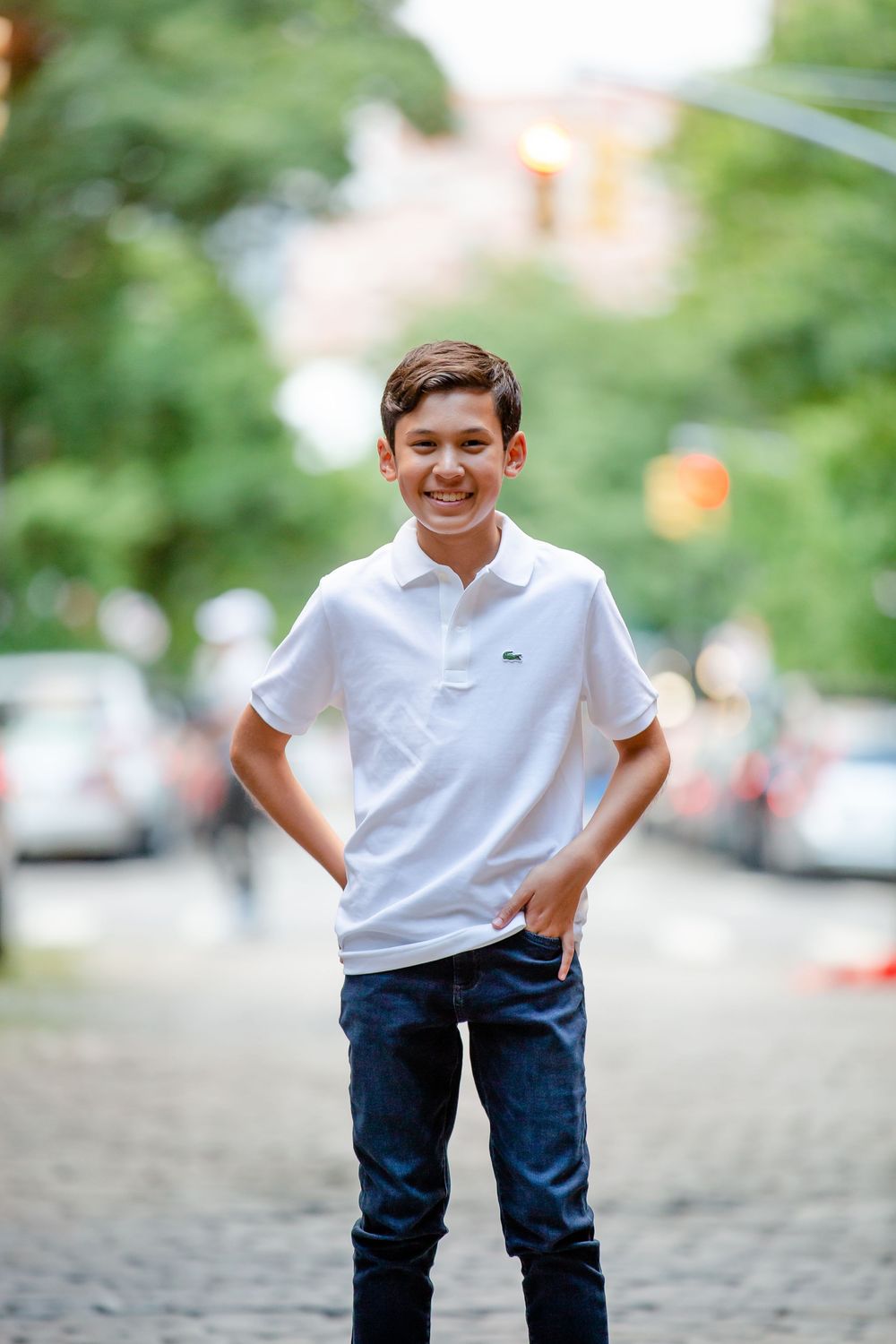 Child in white polo shirt and jeans plays on a cobblestone street with blurred green background.