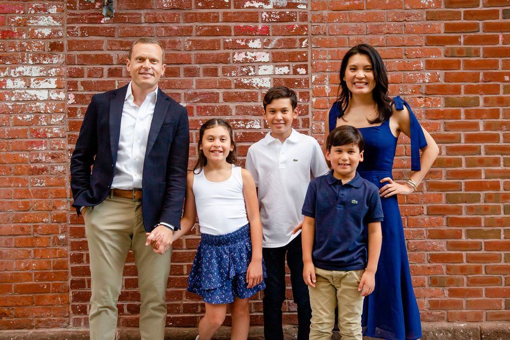 A family of five stands together for a portrait against a rustic red brick wall.