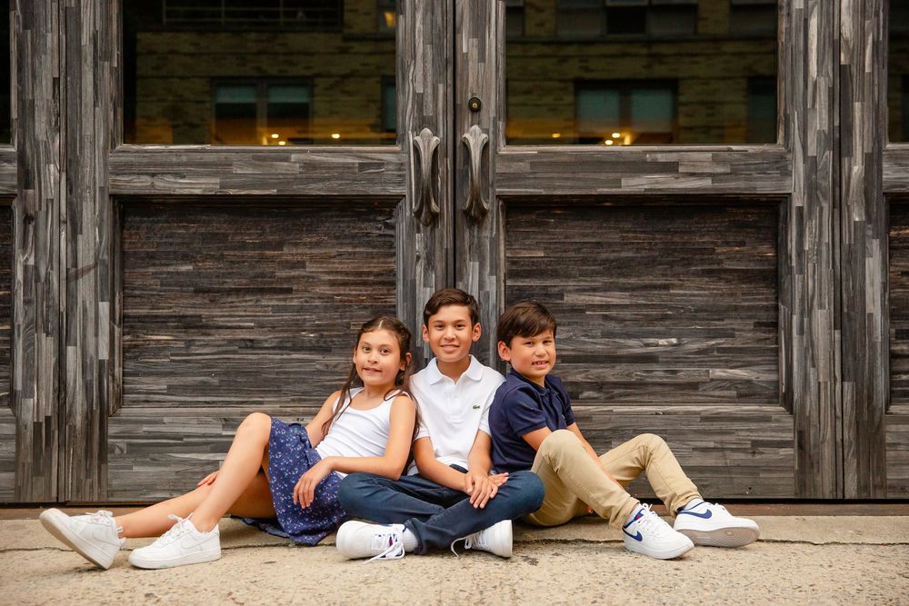 Three children sitting casually against weathered wooden wall outdoors.