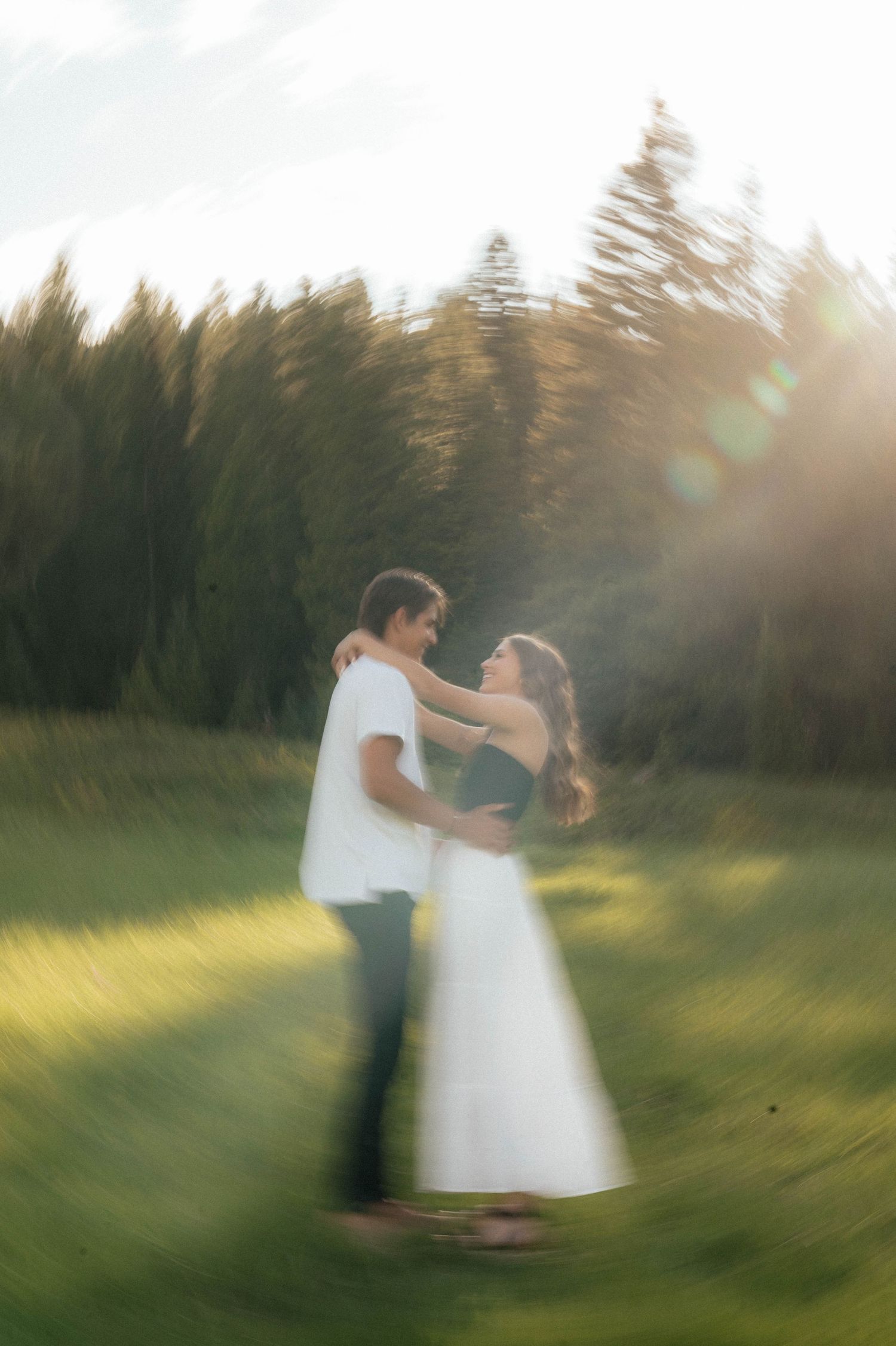 Couple embracing in sunlit green meadow with pine trees, captured with dreamy motion blur effect.