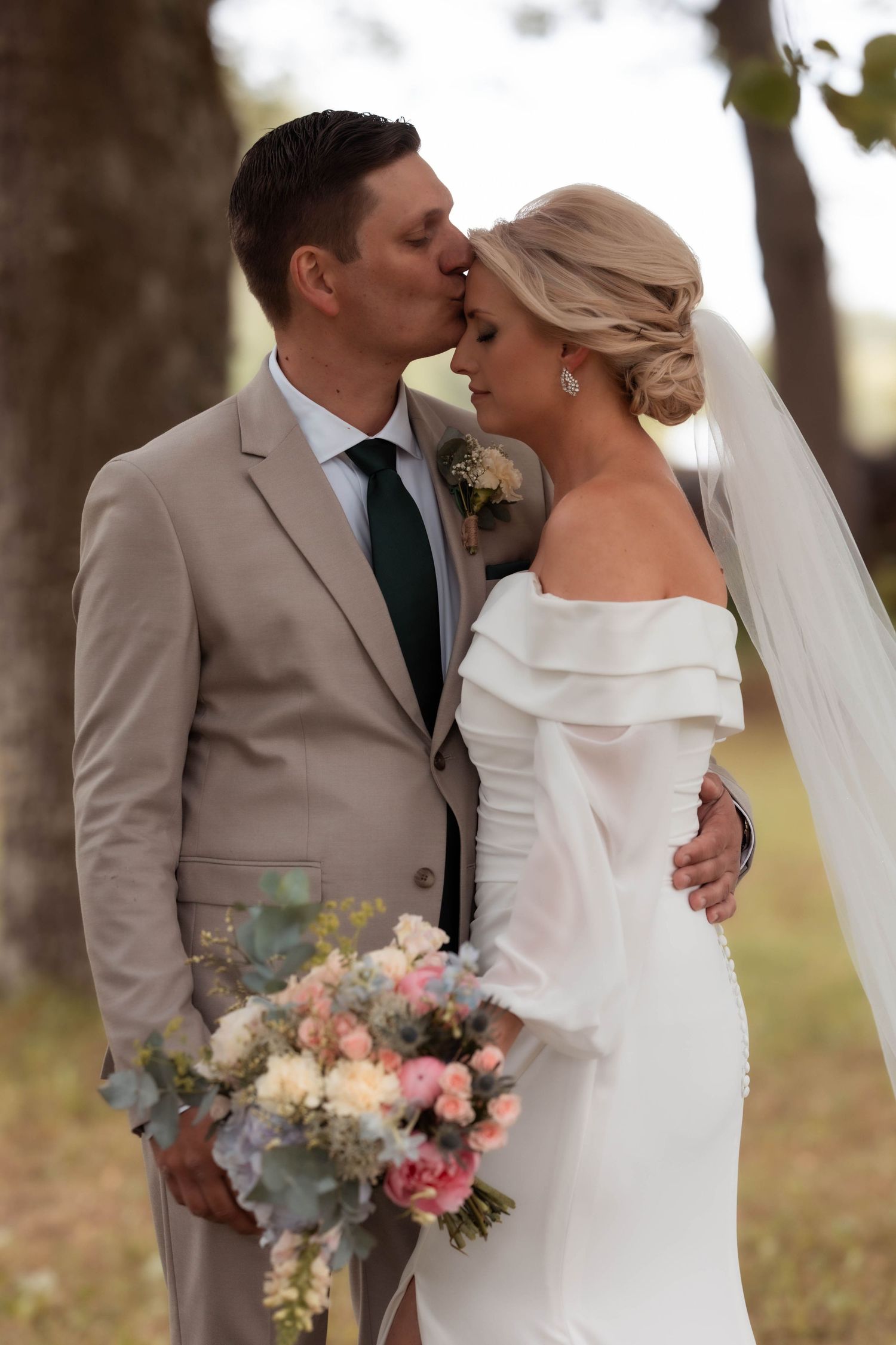 Groom in beige suit kisses bride in off-shoulder white gown holding pastel bouquet outdoors.