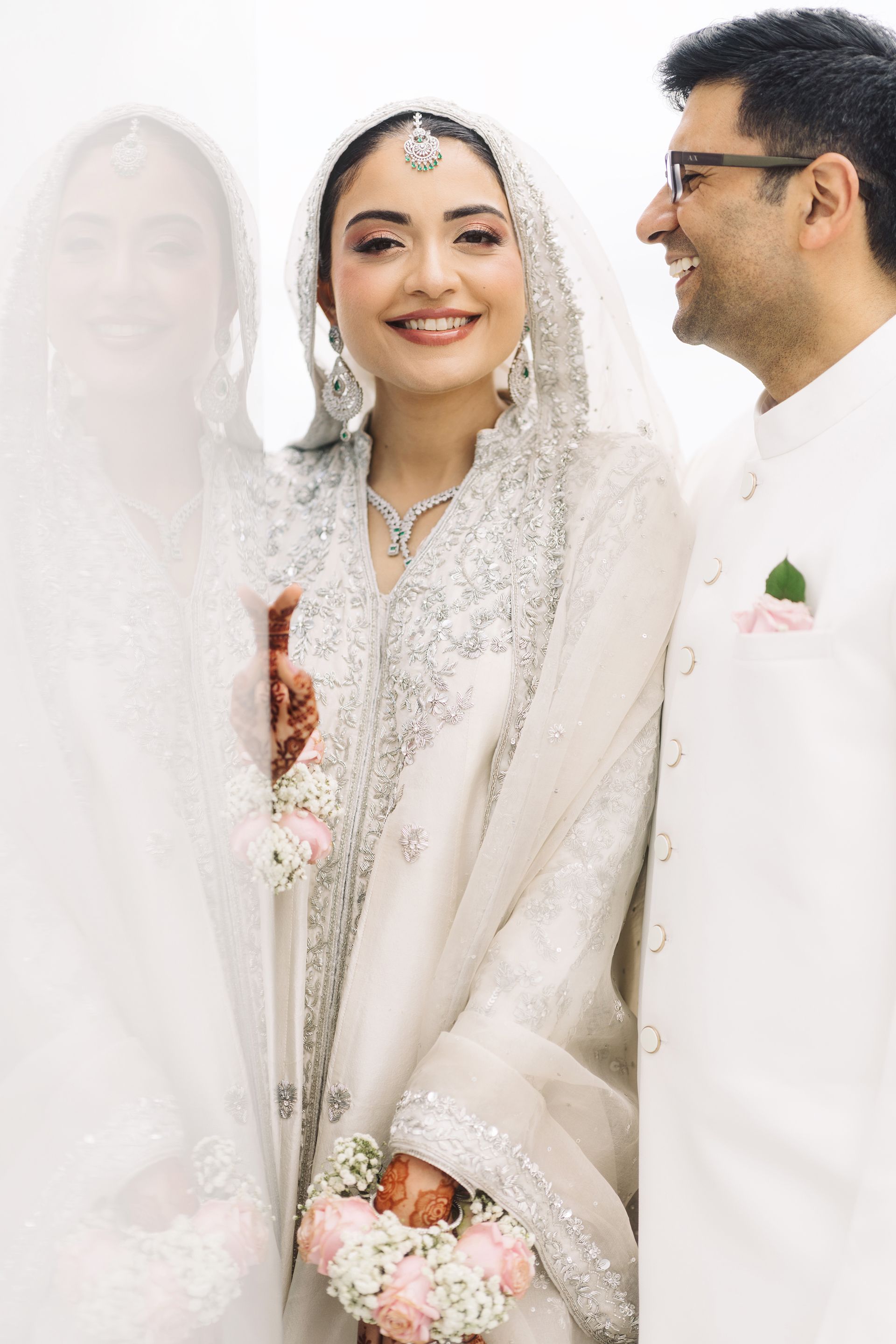 A smiling South Asian bride in white embroidered bridal wear with veil and groom in white sherwani at a wedding ceremony.