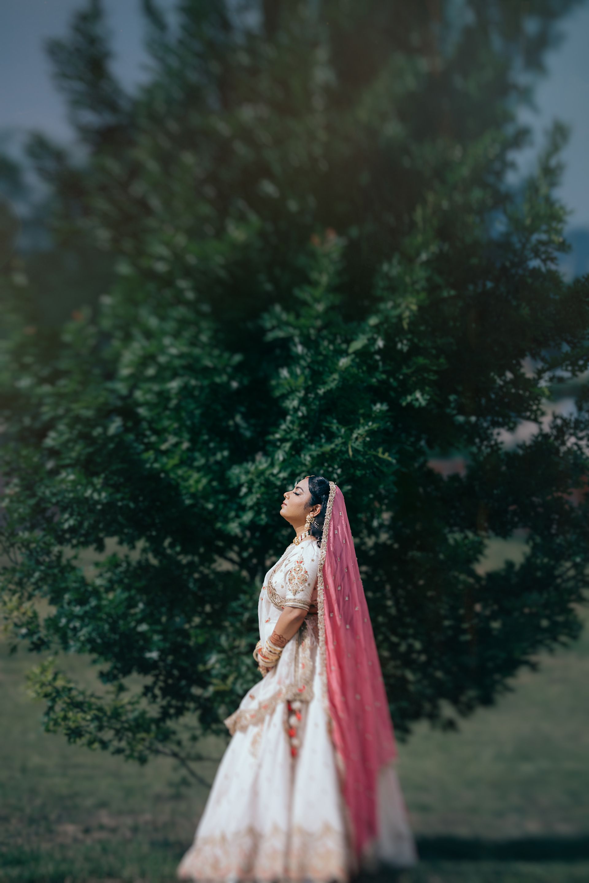 A bride in a white and gold lehenga with a pink dupatta stands gracefully outdoors before a lush green tree.