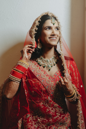 Traditional Indian bride in Houston in red embroidered lehenga and jewelry adjusts her outfit in anelegant pose.