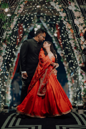 A couple in traditional Indian wedding attire dance together under a romantic floral arch with twinkling lights.