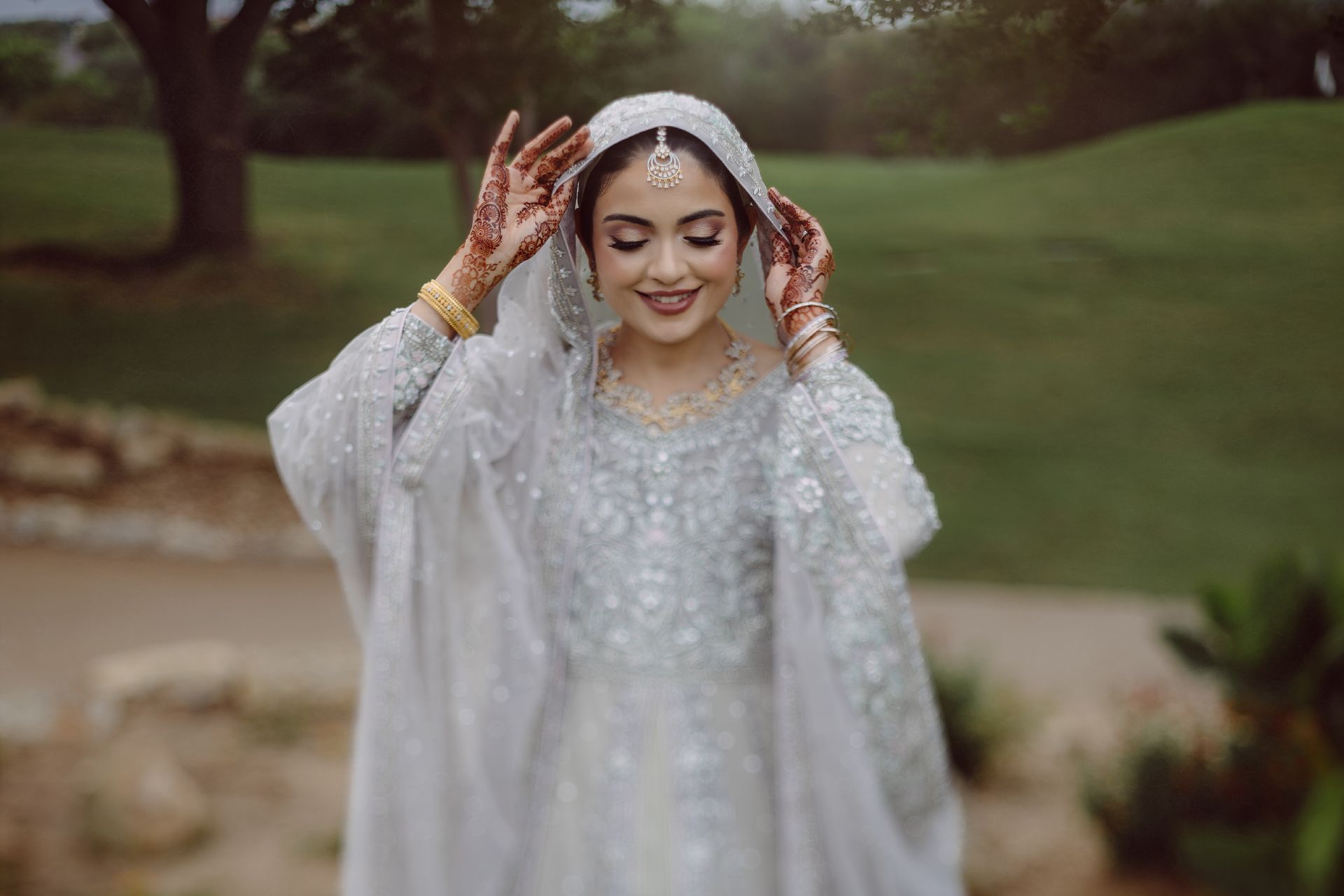 South Asian bride in white embellished lehenga and dupatta with mehndi hands, smiling outdoors.