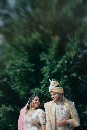Indian bride and groom in traditional wedding attire smile at each other outdoors against lush green foliage backdrop.