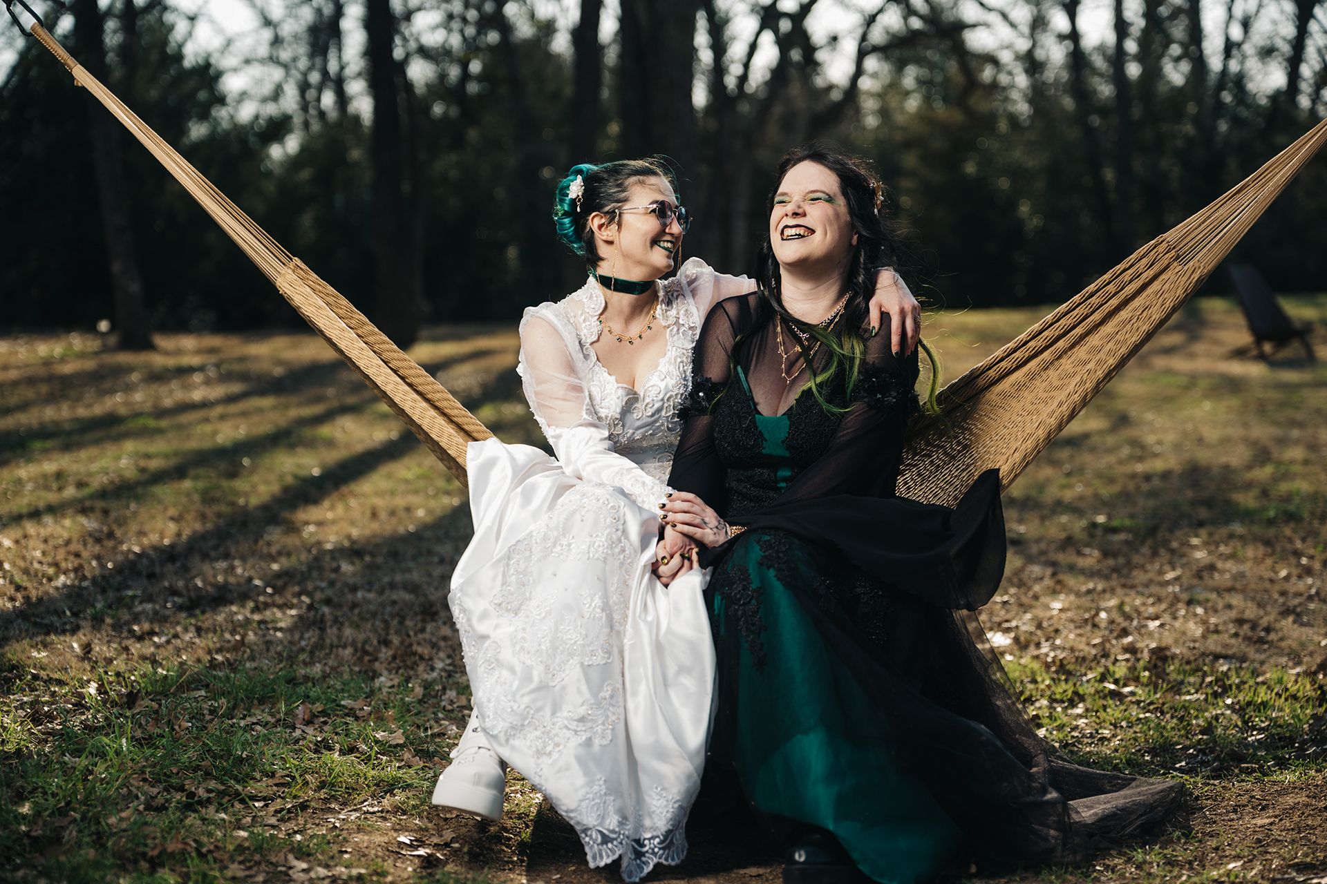 Two brides in wedding gowns laugh joyfully while sitting together in a hammock outdoors.