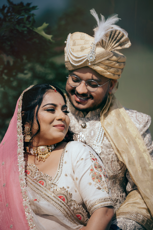 Indian bride and groom in traditional wedding attire, she in white lehenga with pink dupatta, he in gold sherwani and turban.