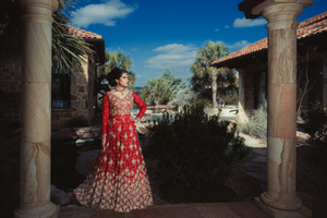A Pakistani bride in a red floral dress poses between stone columns in a Mediterranean-style courtyard setting in Austin, TX.