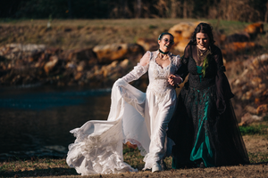 Two people in elegant gowns, one white and one emerald green, walk together by a waterfront at sunset.