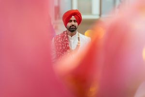 Groom wearing a red turban in a Gurudwara is photographed through soft pink blurred foreground elements.