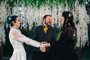 A wedding ceremony takes place under hanging white wisteria flowers with an officiant in a dark suit and yellow tie.
