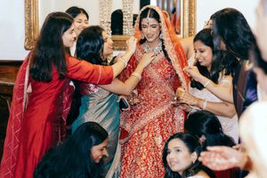 A bride in Houston in a traditional red lehenga is surrounded by friends and family helping her prepare for the wedding ceremony.