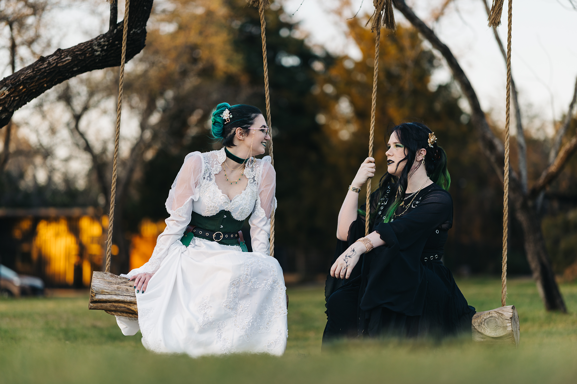 Two women in gothic bridal gowns sit on rope swings outdoors, one in white and one in black, surrounded by trees.
