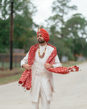 Traditional Sikh groom poses in white attire with red turban and shawl during outdoor wedding photoshoot.