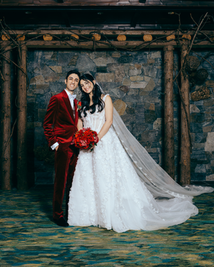 Lake Placid couple in wedding attire poses under a rustic wooden pergola against a stone wall, with a flowing white dress and red suit.