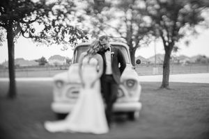 A romantic black and white wedding photo showing a couple with their vintage car under dreamy trees.