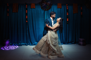 A couple in formal attire dances together during a wedding reception against a blue backdrop with string lights.