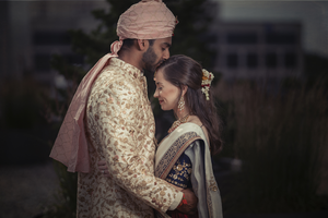 American bride and Indian groom in traditional Indian wedding attire embrace intimately in a moody outdoor setting.