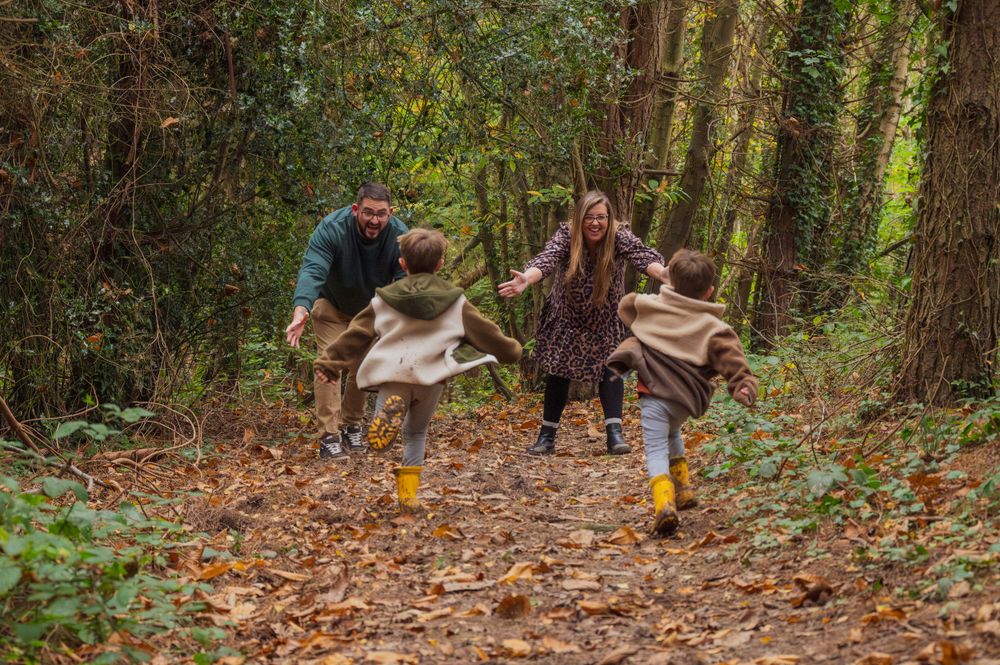 Two people jumping playfully on a leaf-covered forest path while wearing matching yellow boots.