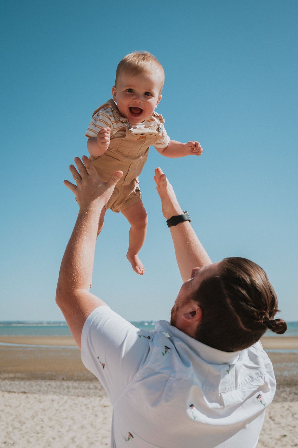 A playful moment at the beach with a small child being lifted high into the air against a bright blue sky.