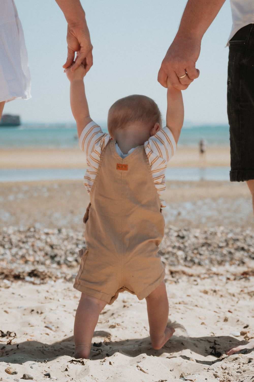 A toddler in khaki shorts stands in soft beach sand against a turquoise ocean backdrop.