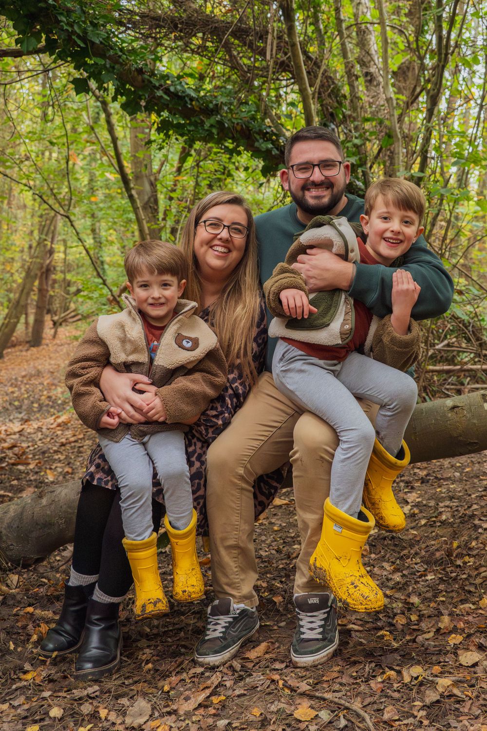 A family sits together on a fallen log in an autumn forest wearing matching yellow boots.