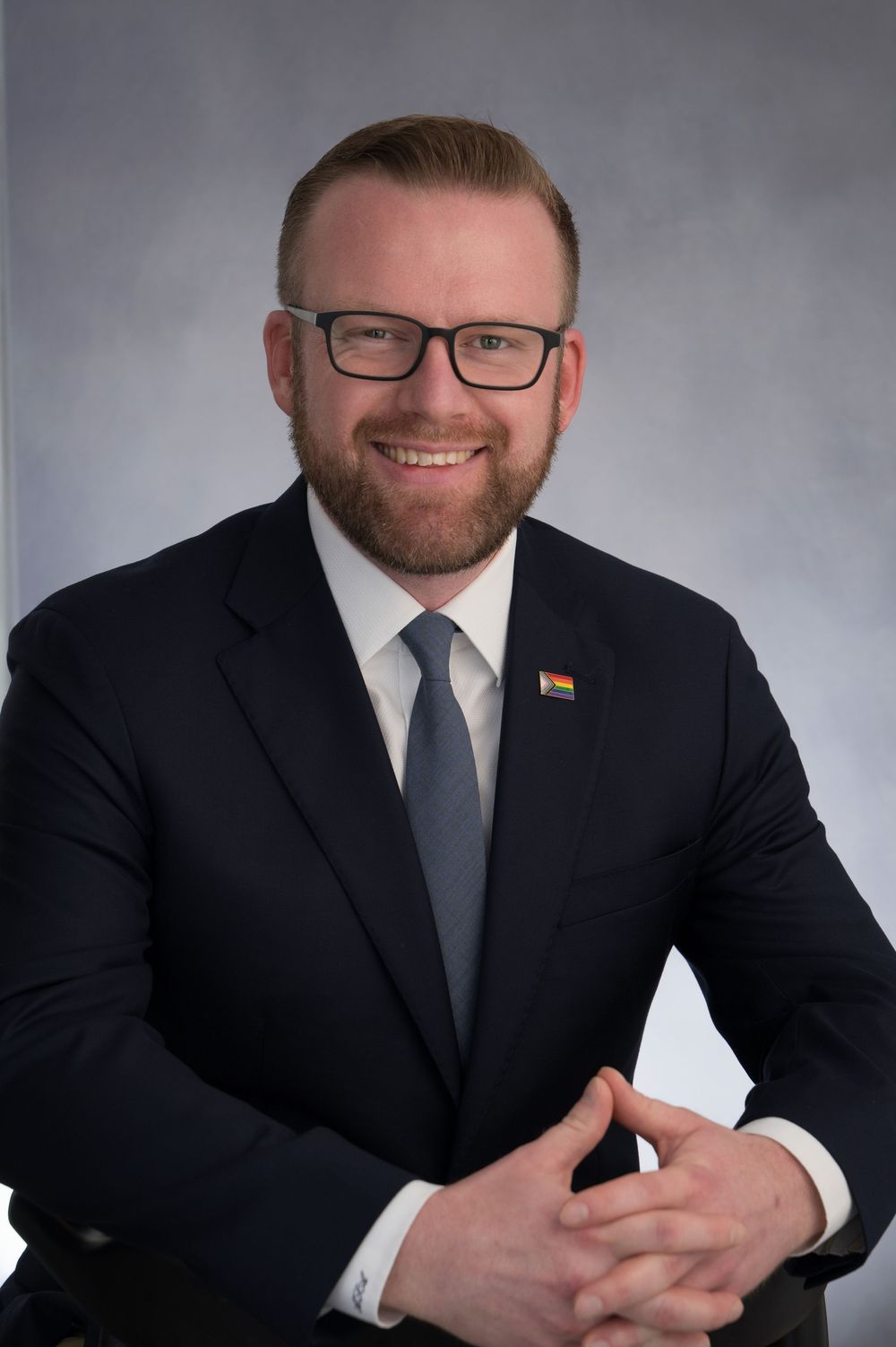 Professional headshot of a smiling bearded man in a dark suit, gray tie, and glasses with a rainbow pin.