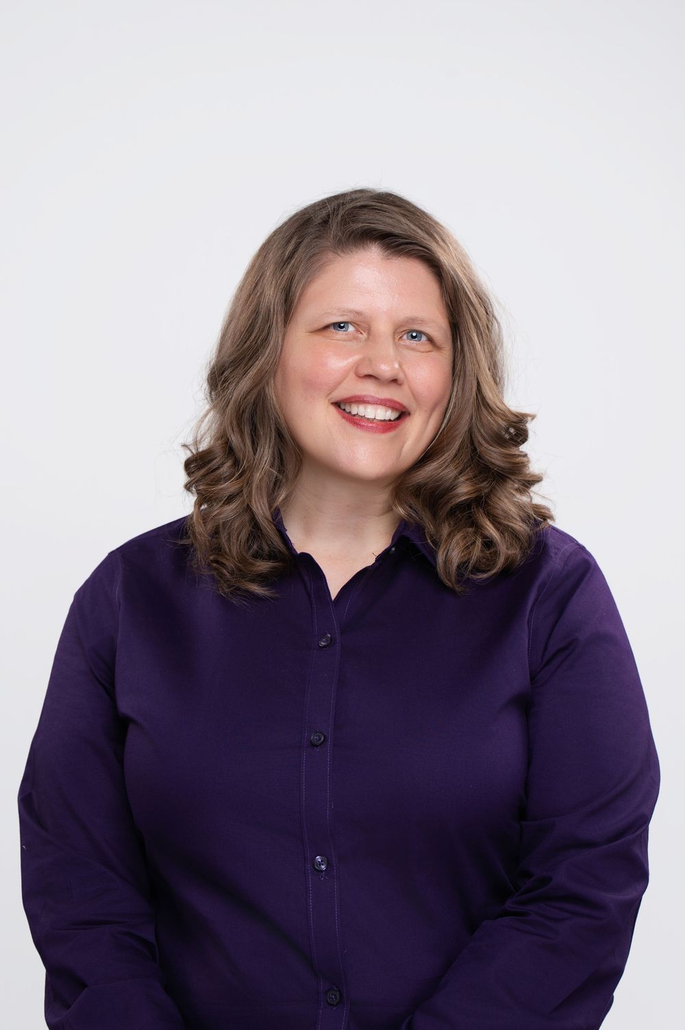 Smiling woman with wavy brown hair wearing a purple button-up blouse against a light gray background.