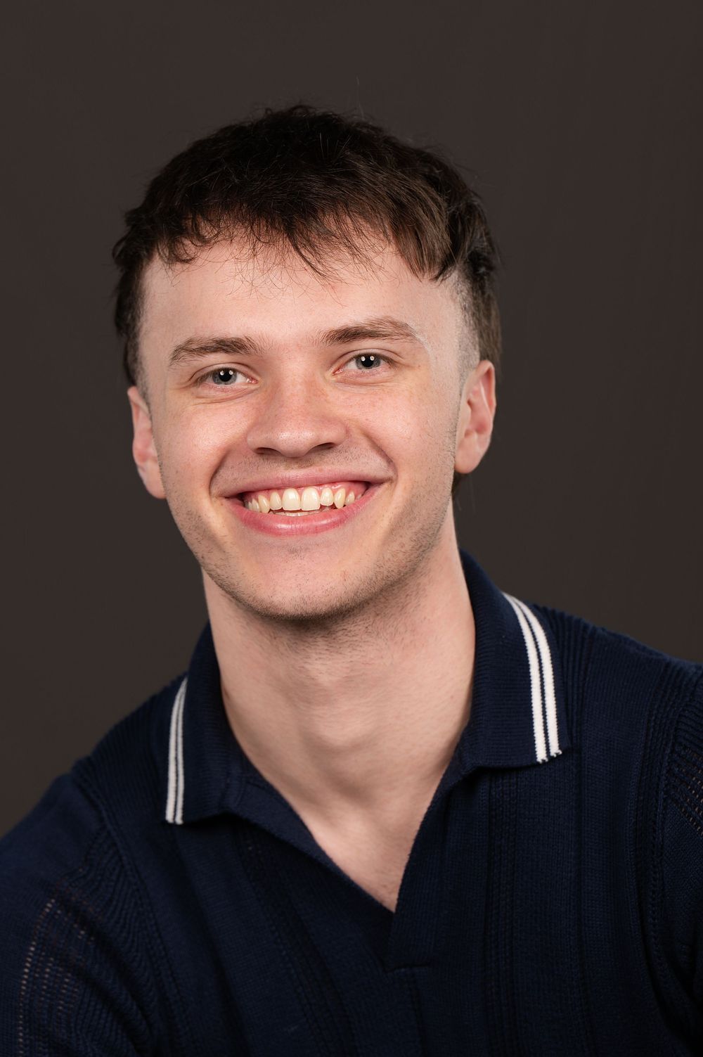 Young man smiling confidently in a navy blue polo shirt against a dark gray background.