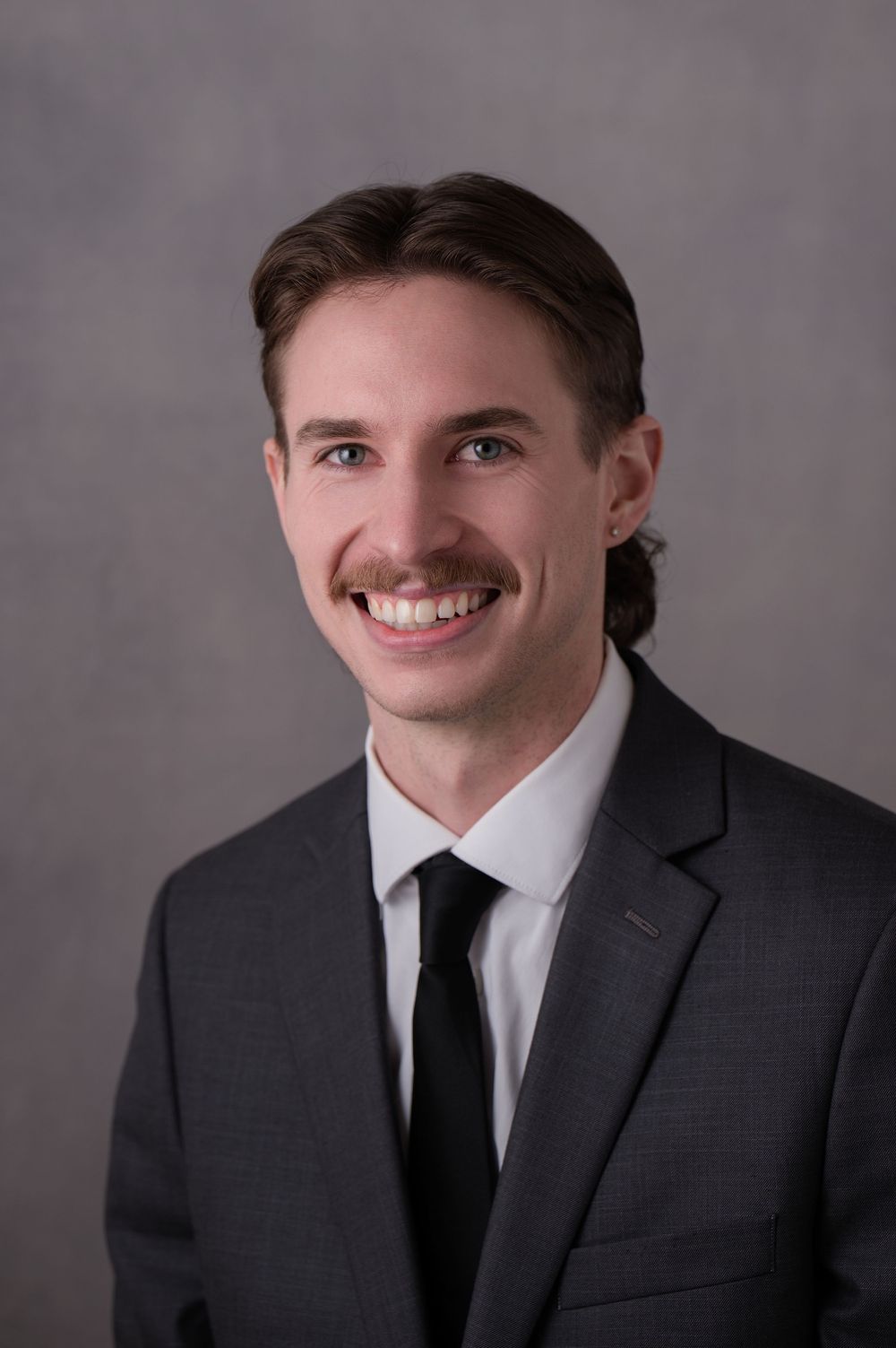 Young professional man with mustache wearing gray suit and black tie smiling in formal headshot photo.