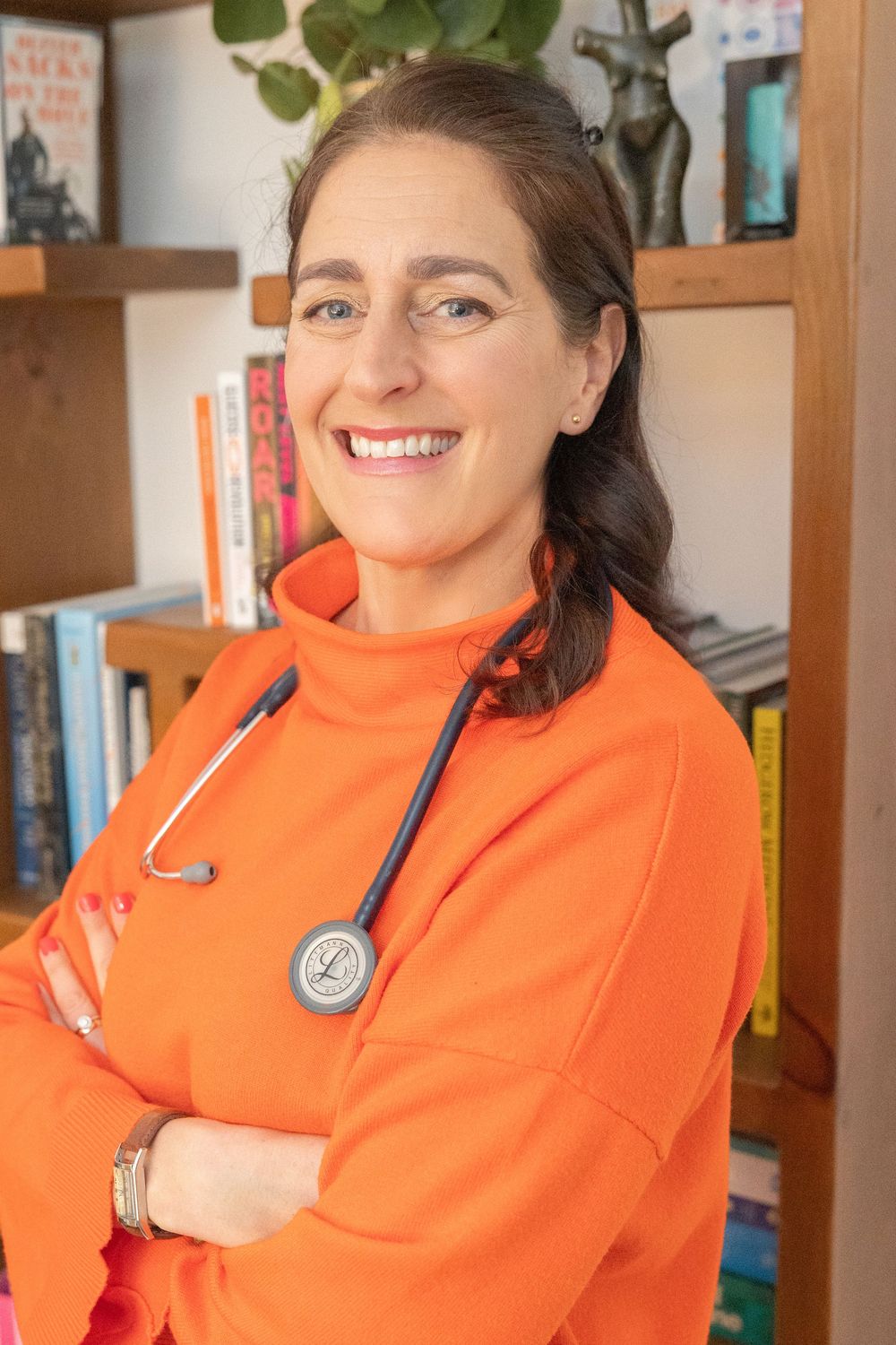 Person wearing a bright orange jacket smiles while sitting in front of a bookshelf in a home setting.