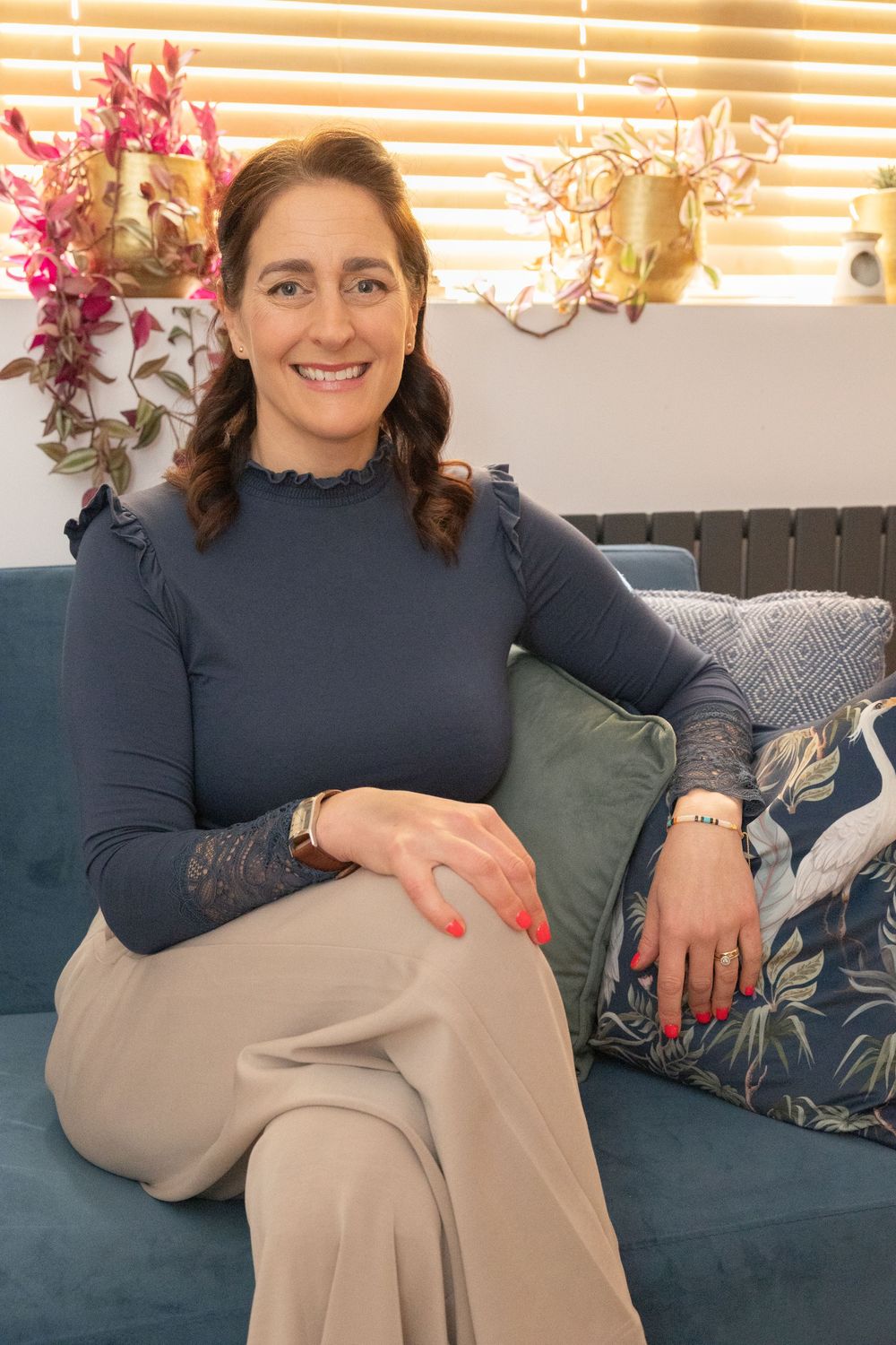 A person wearing a navy sweater and beige pants sits comfortably on a teal couch with floral throw pillows.